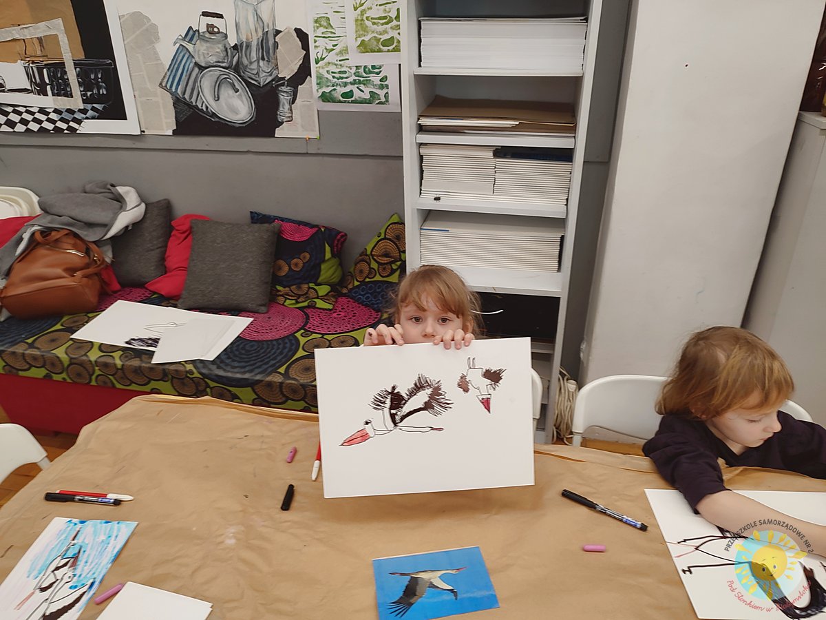 Dziewczynka, siedząca przy stole pokazuje namalowanego bociana - Przedszkole Samorządowe Nr 2 Pod Słonkiem