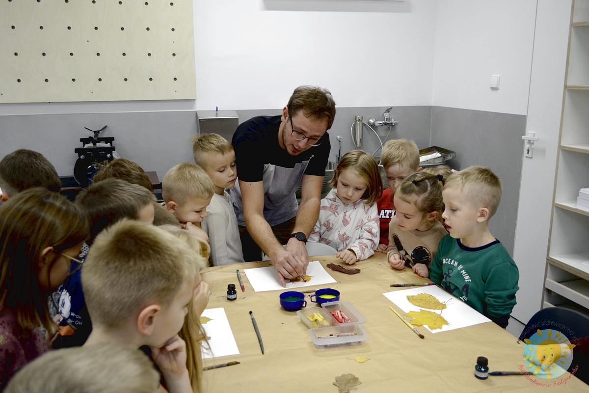 Dzieci stoją obok stołu i obserwują jak nauczyciel maluje liście - Przedszkole Samorządowe Nr 2 Pod Słonkiem w Białymstoku