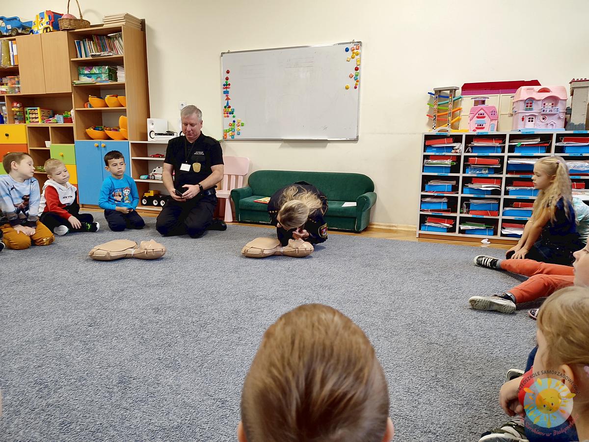 Dzieci siedzą na dywanie a pani wraz z panem ze Straży Miejskiej uczą ich udzielania pierwszej pomocy - Przedszkole Samorządowe Nr 2 Pod Słonkiem w Białymstoku