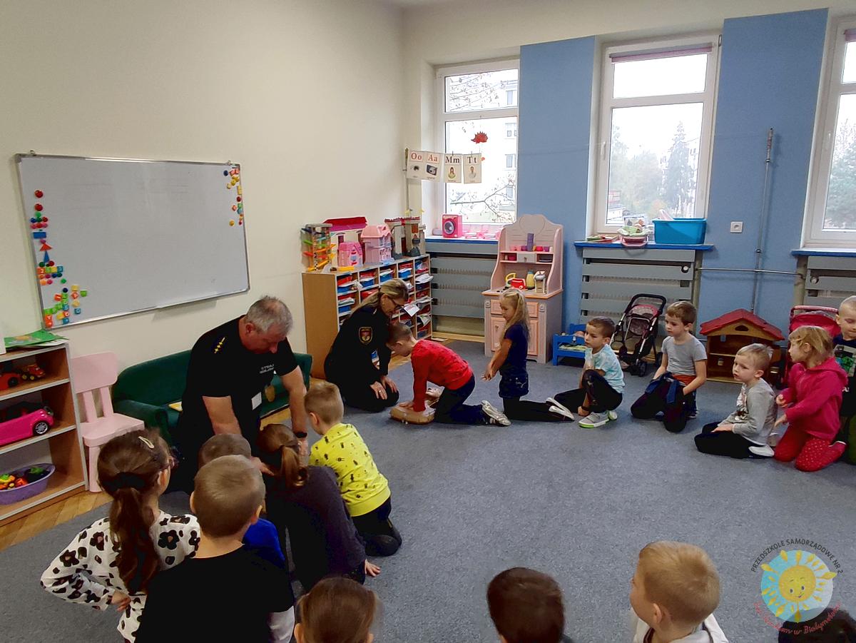 Dzieci siedzą na dywanie a pani wraz z panem ze Straży Miejskiej uczą ich udzielania pierwszej pomocy - Przedszkole Samorządowe Nr 2 Pod Słonkiem w Białymstoku