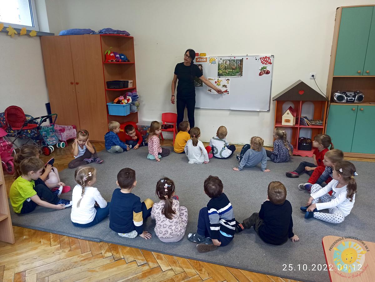 Pani stojąca przy tablicy, tłumaczy dzieciom siedzącym na dywanie - Przedszkole Samorządowe Nr 2 Pod Słonkiem w Białymstoku