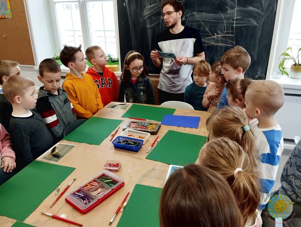 Dzieci siedzą przy stole w galerii Sleńdzińskich i wsłuchują się w komentarz nauczyciela - Przedszkole Samorządowe Nr 2 Pod Słonkiem w Białymstoku