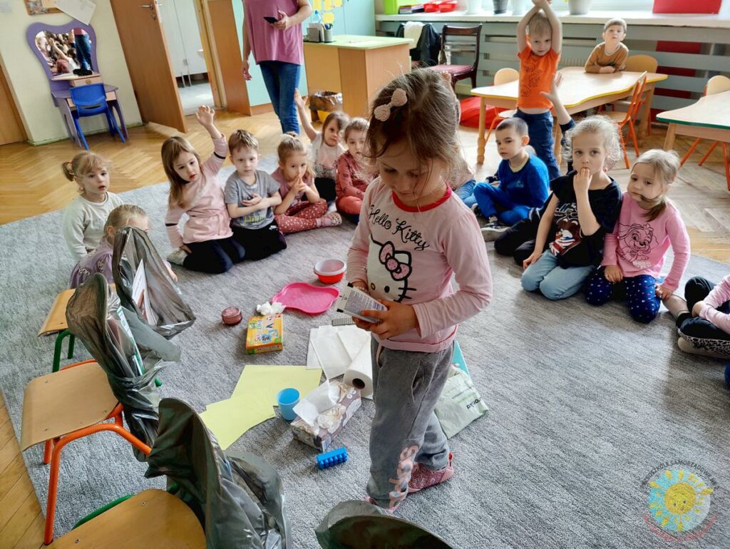 Dzieci siedzą na dywanie i segregują śmieci do worków zawieszonych na krzesłach - Przedszkole Samorządowe Nr 2 Pod Słonkiem w Białymstoku