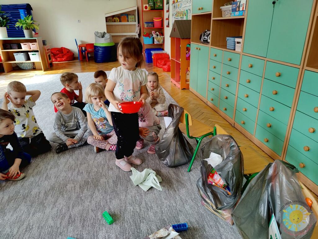 Dzieci siedzą na dywanie i segregują śmieci do worków zawieszonych na krzesłach - Przedszkole Samorządowe Nr 2 Pod Słonkiem w Białymstoku