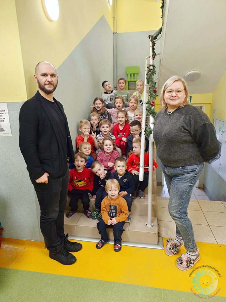 Dzieci w wieku przedszkolnym siedzą na schodach a obok stoi mężczyzna i kobieta - Przedszkole Samorządowe Nr 2 Pod Słonkiem w Białymstoku