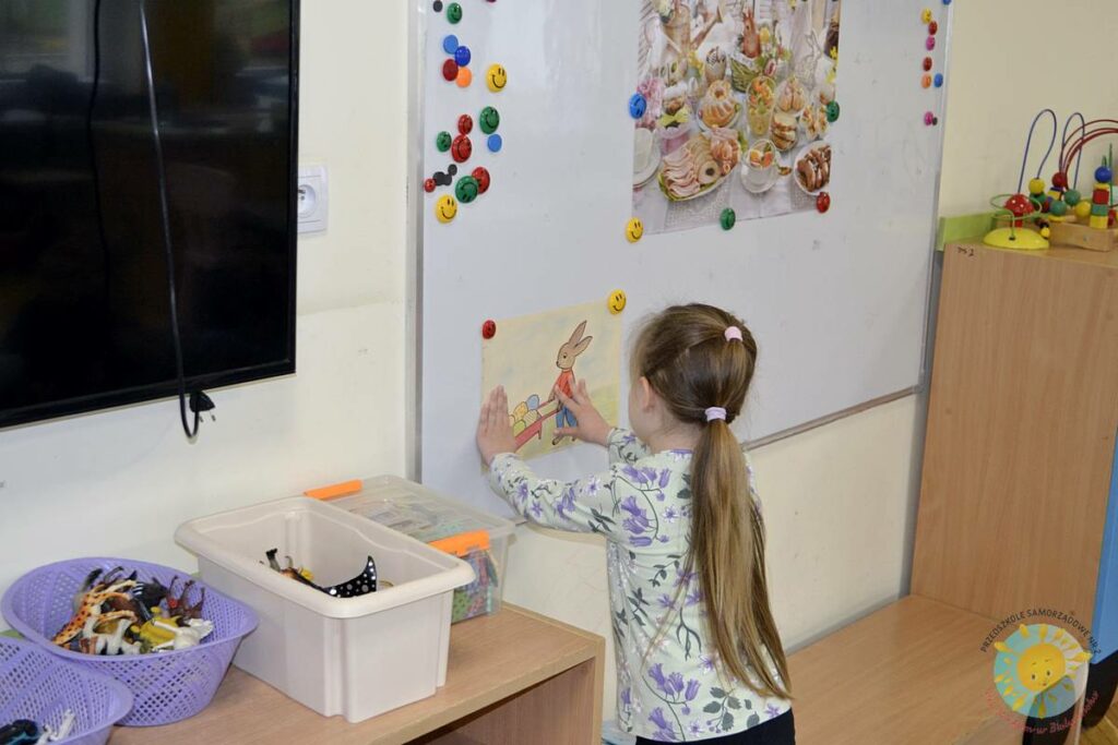 Stojące dziecko przy tablicy, wieszające obrazek - Przedszkole Samorządowe Nr 2 Pod Słonkiem w Białymstoku