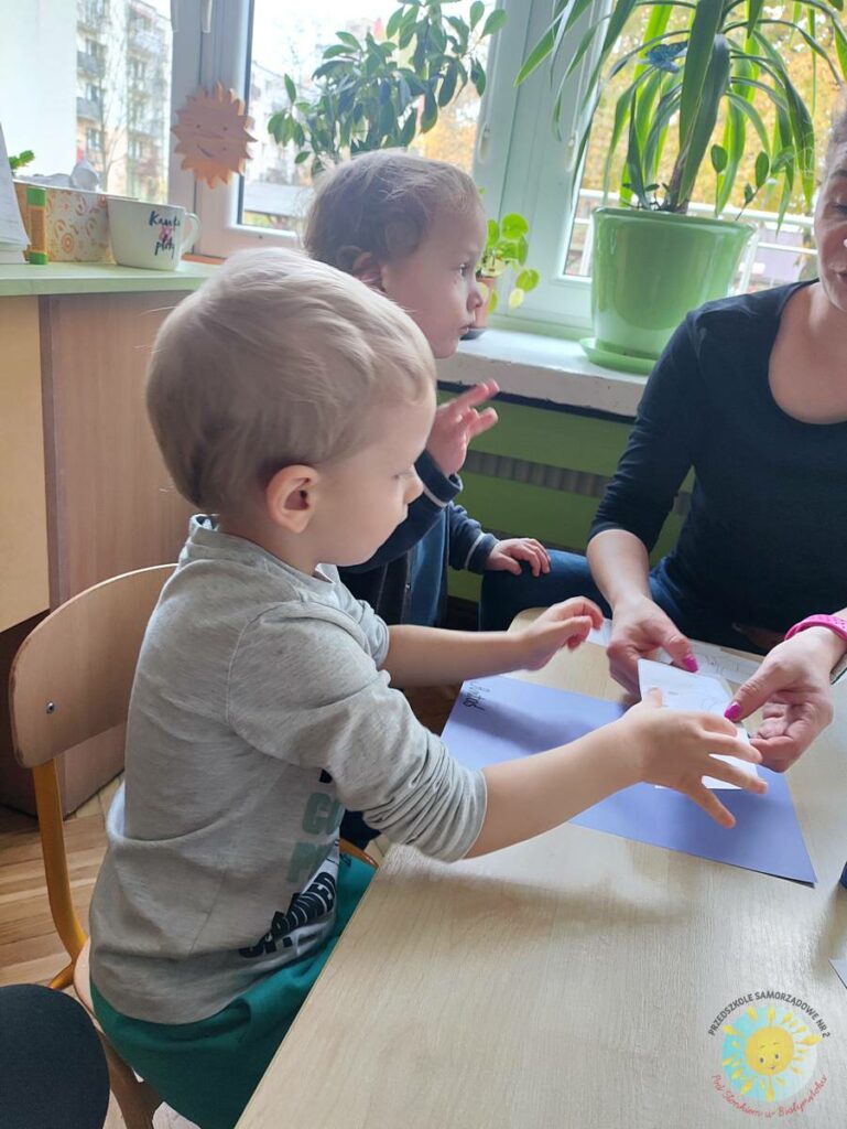 Siedzące dzieci przy przedszkolnym stoliku, wykonują pracę - Przedszkole Samorządowe Nr 2 Pod Słonkiem w Białymstoku