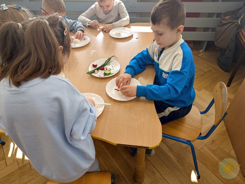 Siedzące dzieci przy stole, które biorą udział w zajęciach gastronomicznych - Przedszkole Samorządowe Nr 2 Pod Słonkiem w Białymstoku