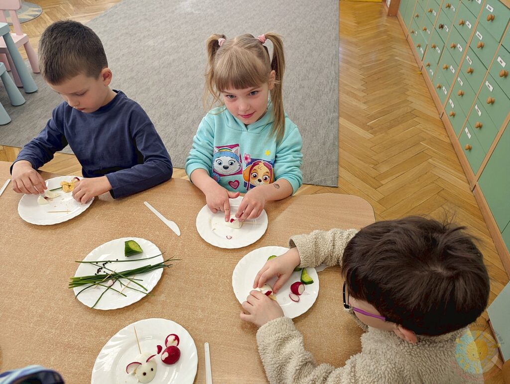 Siedzące dzieci przy stole, które biorą udział w zajęciach gastronomicznych - Przedszkole Samorządowe Nr 2 Pod Słonkiem w Białymstoku