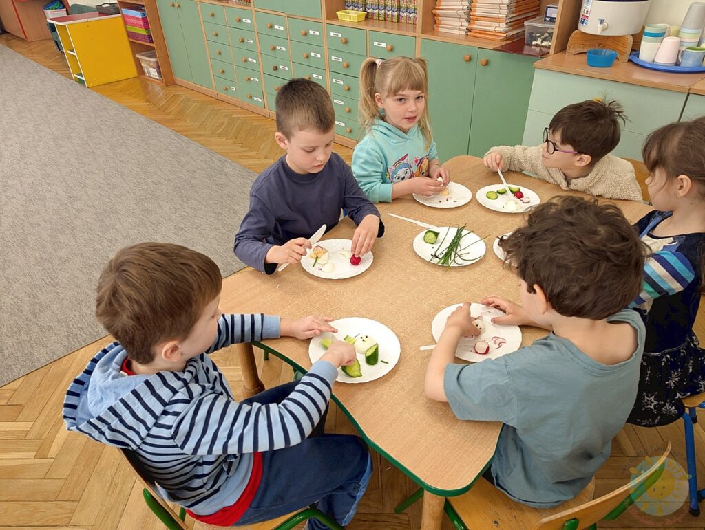 Siedzące dzieci przy stole, które biorą udział w zajęciach gastronomicznych - Przedszkole Samorządowe Nr 2 Pod Słonkiem w Białymstoku