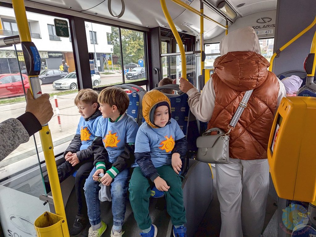 Dzieci w jasnoniebieskich koszulkach z nadrukiem słońca siedzą na siedzeniach w autobusie miejskim, jadąc na wycieczkę.