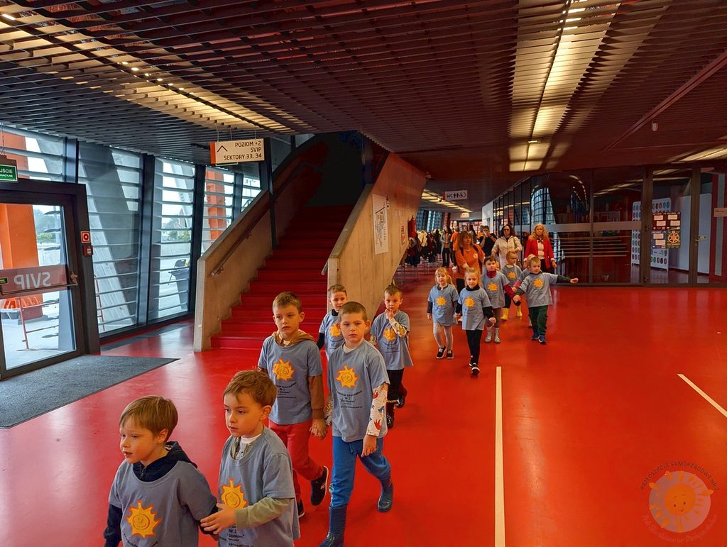 Dzieci w jednolitych niebieskich koszulkach z nadrukiem słońca idą parami przez nowoczesny, przestronny korytarz z czerwoną podłogą na stadionie.