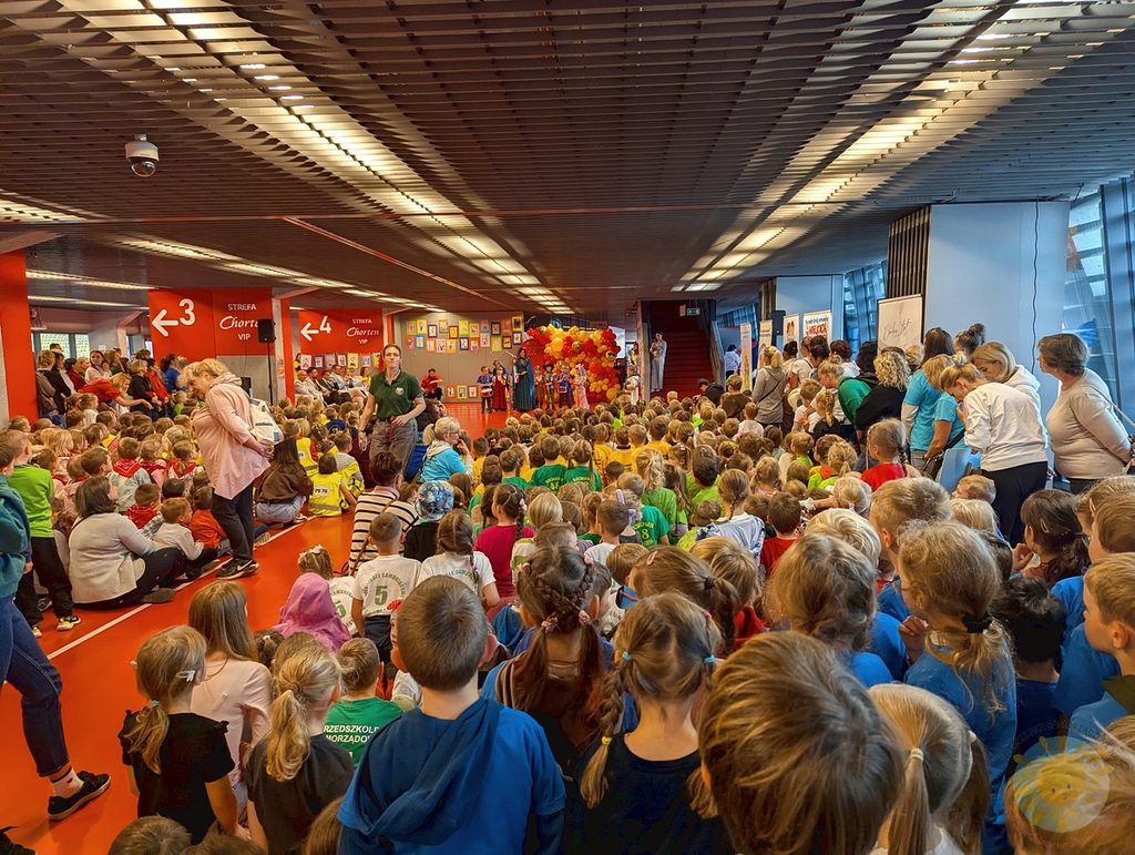 Duża grupa dzieci siedzących w hali sportowej podczas uroczystości, zwróconych w stronę dekoracji z balonów i prowadzącego.