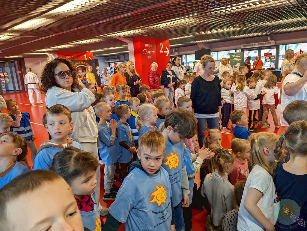 Grupa dzieci w błękitnych koszulkach z nadrukiem słońca oraz dorośli opiekunowie uczestniczą w spotkaniu wewnątrz hali sportowej.