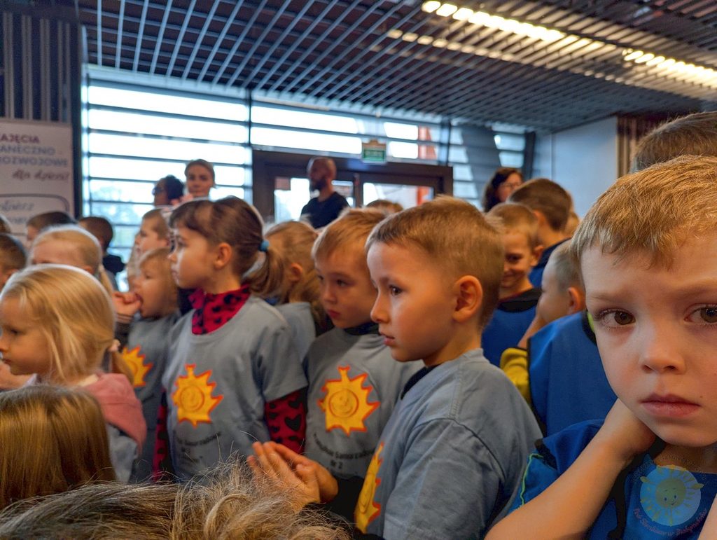 Dzieci w jednakowych niebieskich koszulkach z nadrukiem słońca skupione wewnątrz budynku, patrzą w jednym kierunku podczas obchodów święta.
