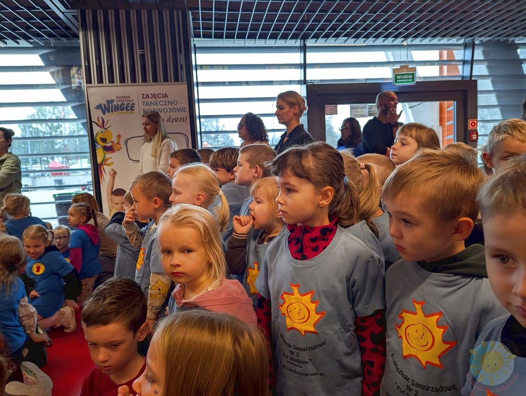 Grupa dzieci w niebieskich koszulkach z logo słońca uczestniczy w wydarzeniu odbywającym się w jasnej sali sportowej.