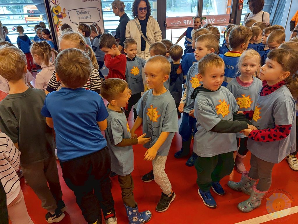 Grupa dzieci w jasnoniebieskich koszulkach z nadrukiem słońca trzyma się za ręce w parach podczas zajęć wewnątrz budynku.