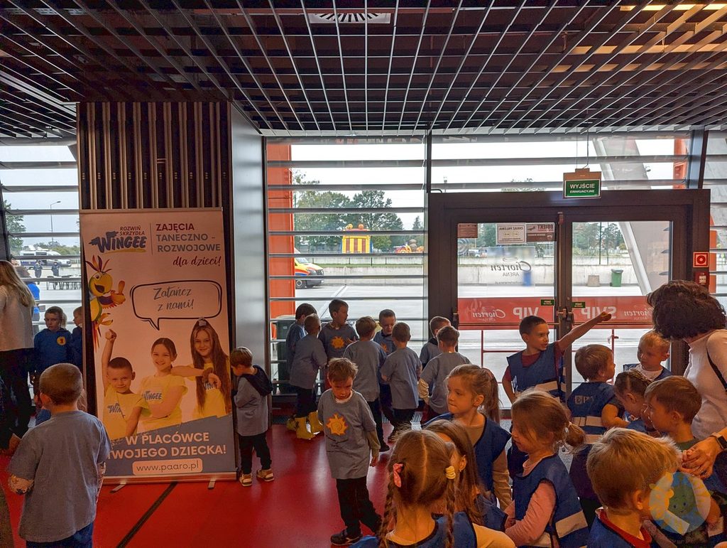 Dzieci w jednakowych koszulkach z nadrukami stoją w hali sportowej obok roll-upu z ofertą zajęć tanecznych dla dzieci.