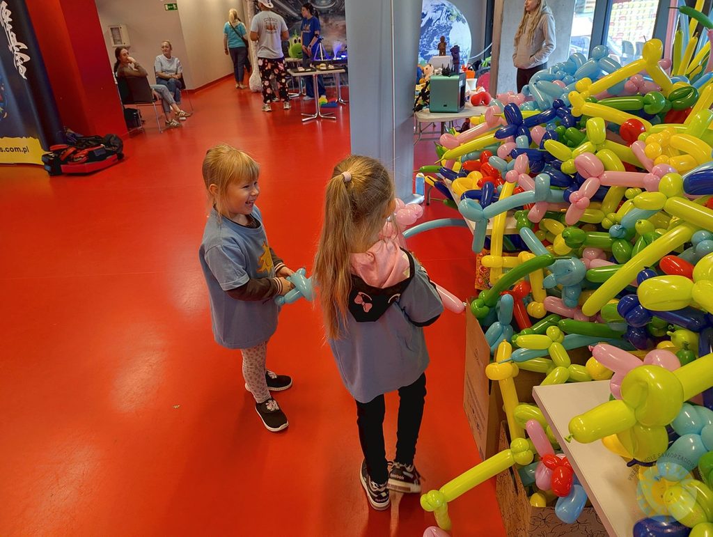 Dzieci przyglądają się dużej stercie kolorowych balonów o różnych kształtach w jasnym pomieszczeniu.