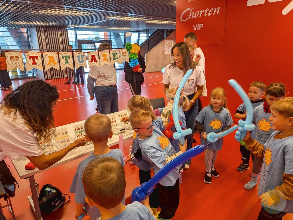 Dzieci w jasnoniebieskich koszulkach z nadrukiem słońca bawią się na stadionie miejskim, korzystając ze stanowiska z tatuażami i balonowymi mieczami.