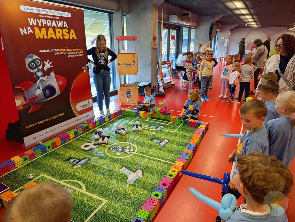 Dzieci obserwują małe roboty grające w piłkę nożną na specjalnej macie, obok ustawiono baner reklamowy warsztatów robotyki „Wyprawa na Marsa”.