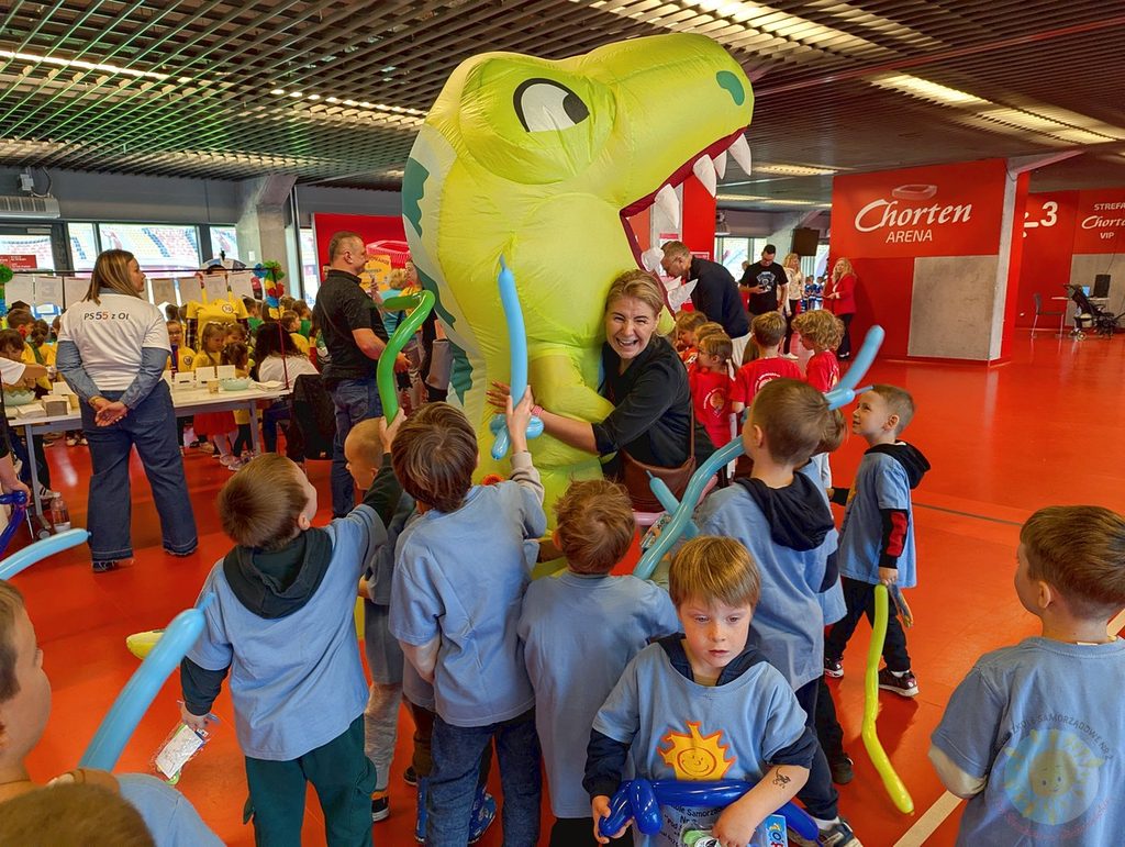 Dzieci w jasnoniebieskich koszulkach otaczają dmuchaną figurę dinozaura, którą przytula uśmiechnięta dorosła osoba.