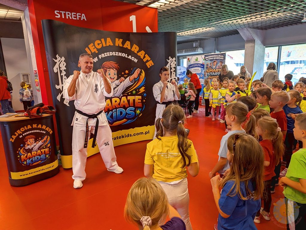 Dwie osoby w strojach do karate prezentują postawy walki przed grupą dzieci na Stadionie Miejskim, w tle widoczne banery z napisem Zajęcia Karate w Przedszkolach.
