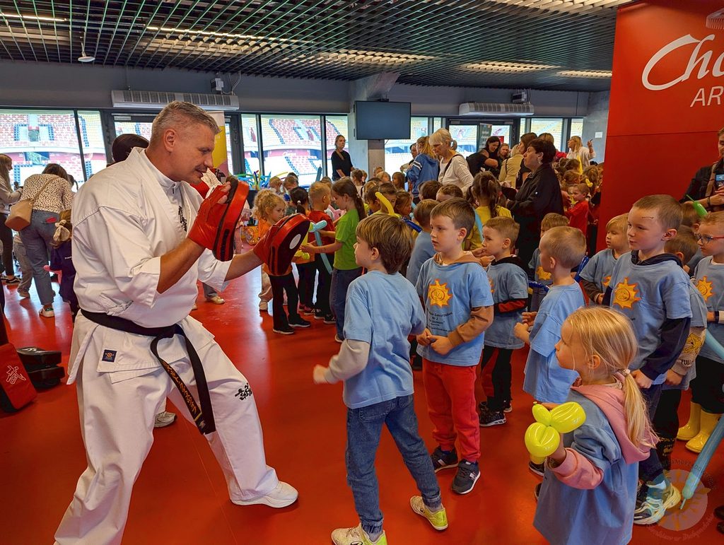 Instruktor w białym stroju do karate prezentuje techniki uderzeń w tarcze, przed nim stoi grupa dzieci z przedszkola obserwująca pokaz.