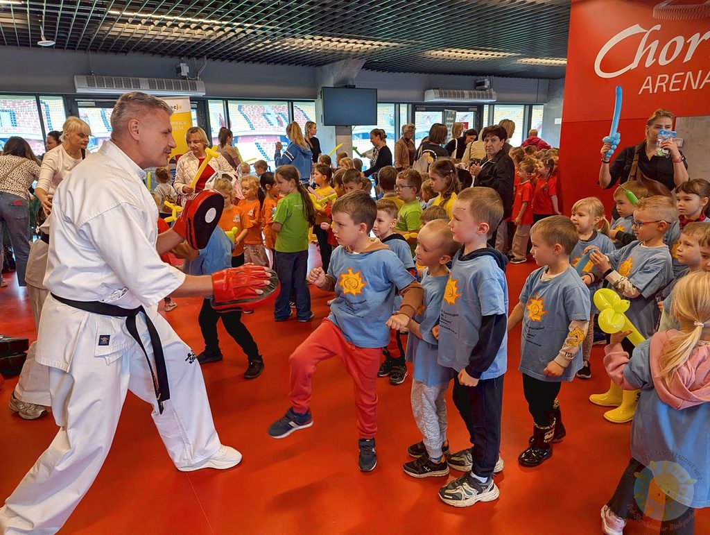 Instruktor w stroju do karate pokazuje dziecku technikę kopnięcia w tarczę treningową w otoczeniu innych dzieci uczestniczących w zajęciach na hali sportowej.