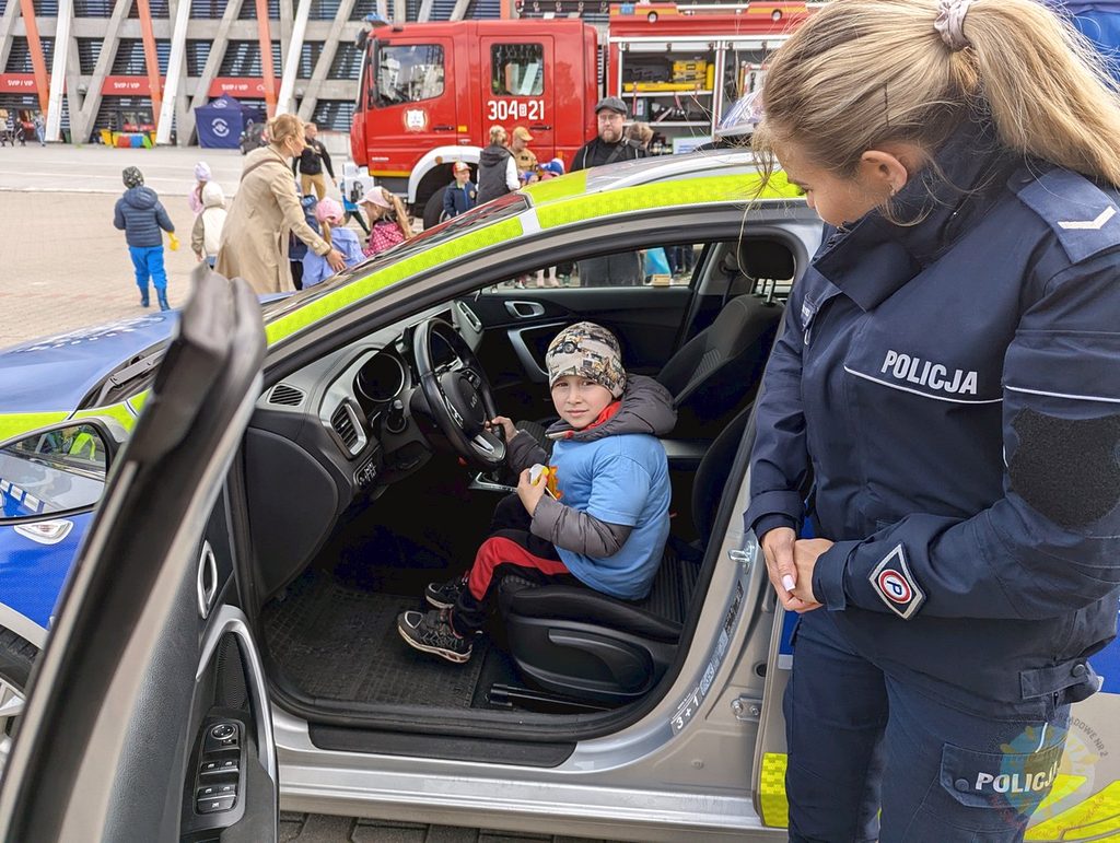 Dziecko siedzi za kierownicą radiowozu, a obok stoi funkcjonariuszka policji. W tle widać wóz strażacki oraz inne dzieci przebywające na stadionie.