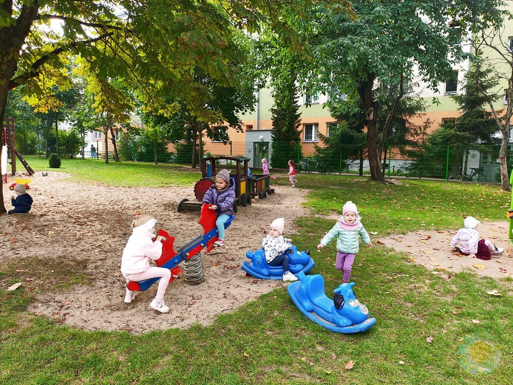 Dzieci bawią się na przedszkolnym placu zabaw, korzystając z huśtawek sprężynowych i drewnianej konstrukcji w kształcie pociągu.