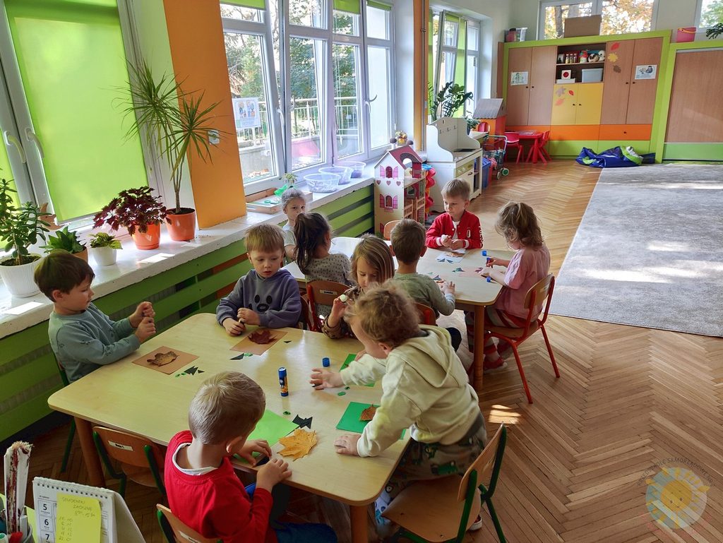 Dzieci siedzące przy stolikach w sali przedszkolnej wykonują prace plastyczne z wykorzystaniem kolorowych liści.