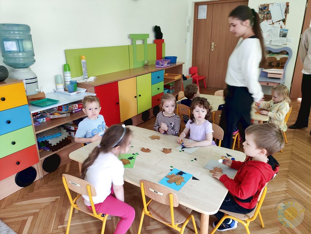Dzieci siedzące przy stolikach w sali przedszkolnej podczas zajęć plastycznych z wykorzystaniem jesiennych liści.