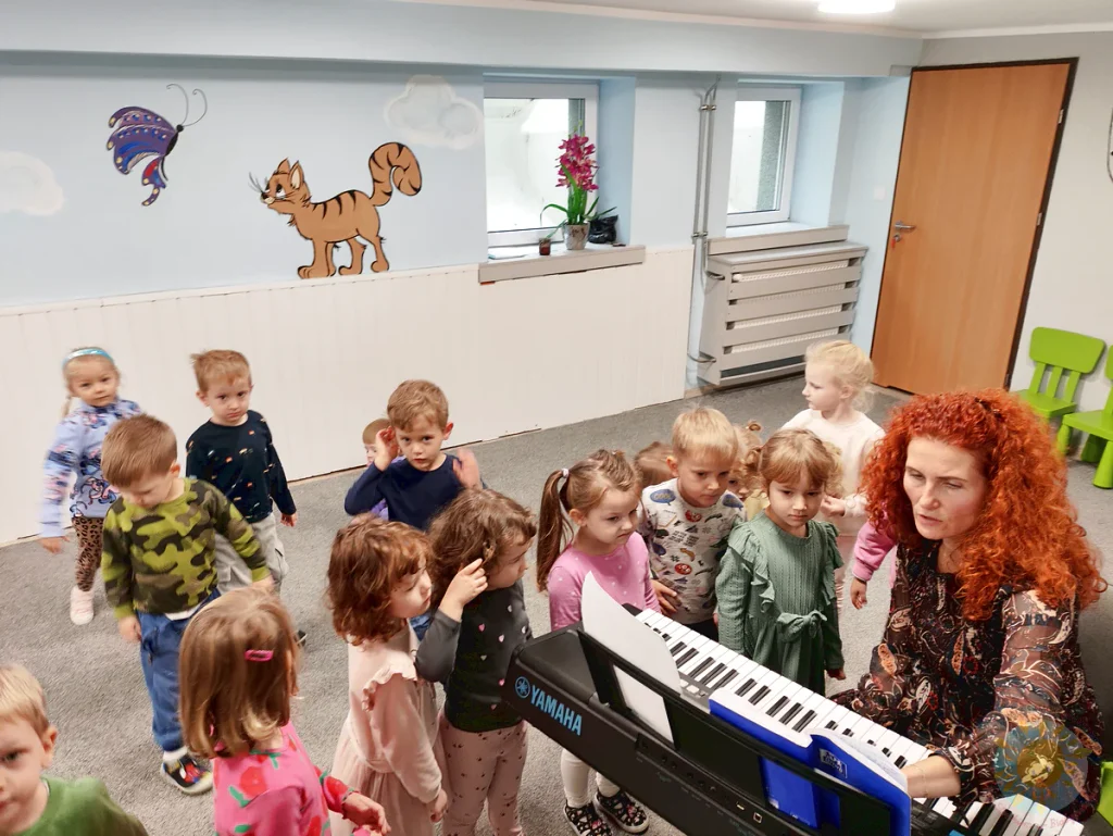 Dorosła osoba gra na pianinie elektronicznym, otoczona przez grupę dzieci uczestniczących w zajęciach muzycznych.