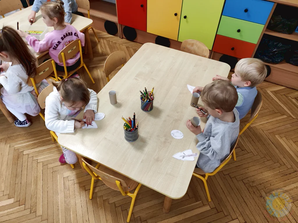 Dzieci siedzące przy stolikach w sali zajęć kolorują wycięte z papieru kształty zwierząt i drzew, korzystając z kredek i rolek papierowych.