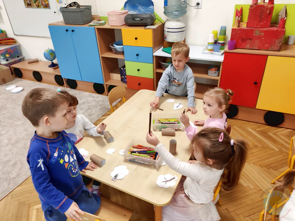 Dzieci siedzące przy stole podczas zajęć plastycznych, kolorujące wycięte z papieru kształty drzew i zwierząt.