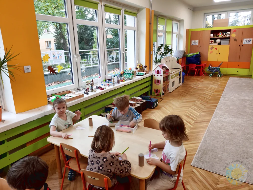 Dzieci siedzą przy stoliku w sali przedszkolnej i wykonują prace plastyczne z wykorzystaniem papierowych elementów.
